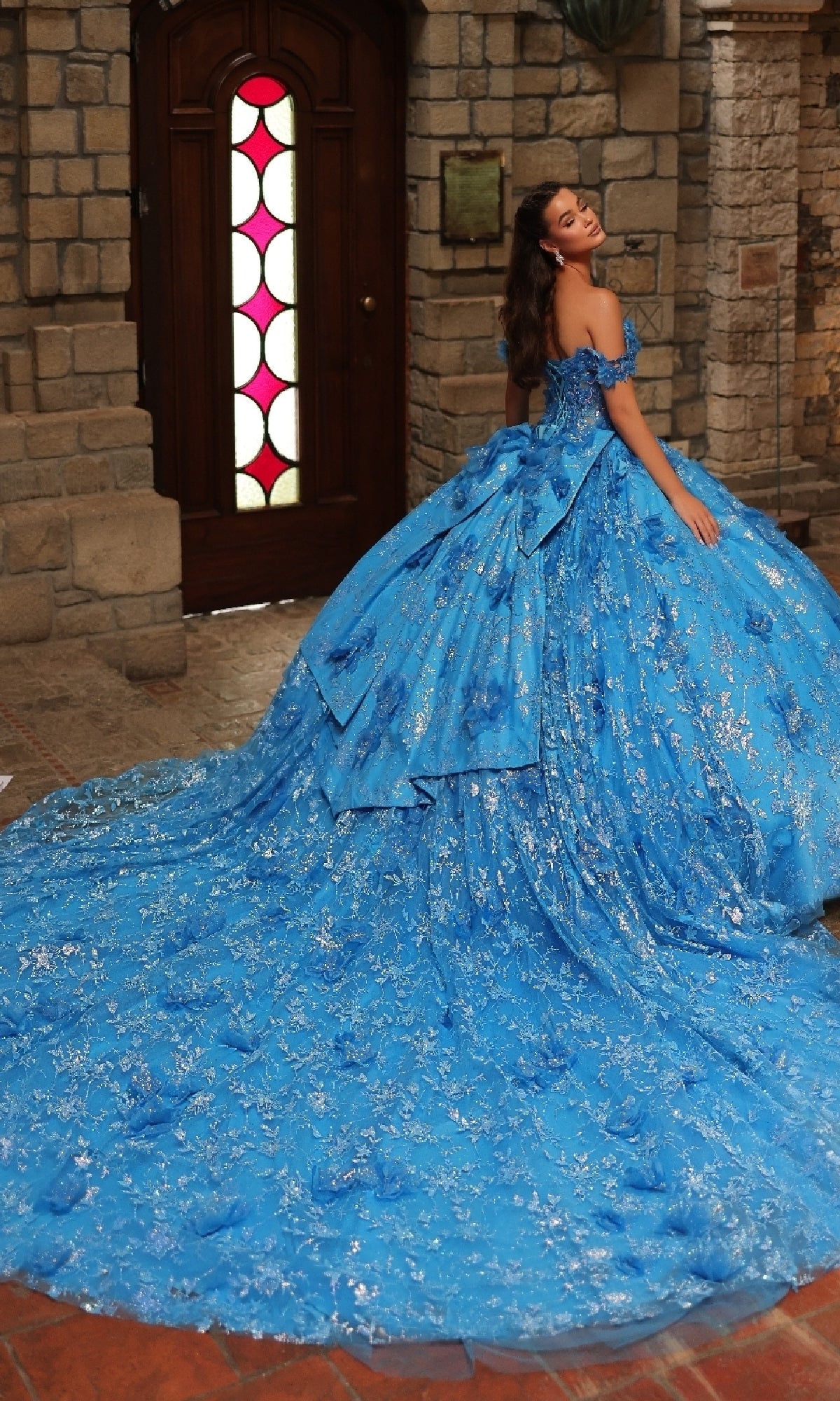 A woman poses indoors in the Amarra 54243 Off-Shoulder Quinceanera Dress, featuring an elaborate blue ball gown with 3D floral appliques, silver accents, and a long train, set against a wooden door and stone walls.